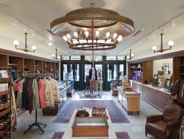 Upscale boutique interior with wooden shelving, clothing racks, an ornate vintage chandelier, patterned rugs, and a leather armchair