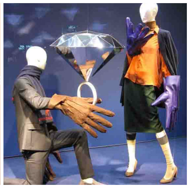 Two faceless mannequins in a blue-toned display, one kneeling offering an oversized ring with a diamond shape overhead