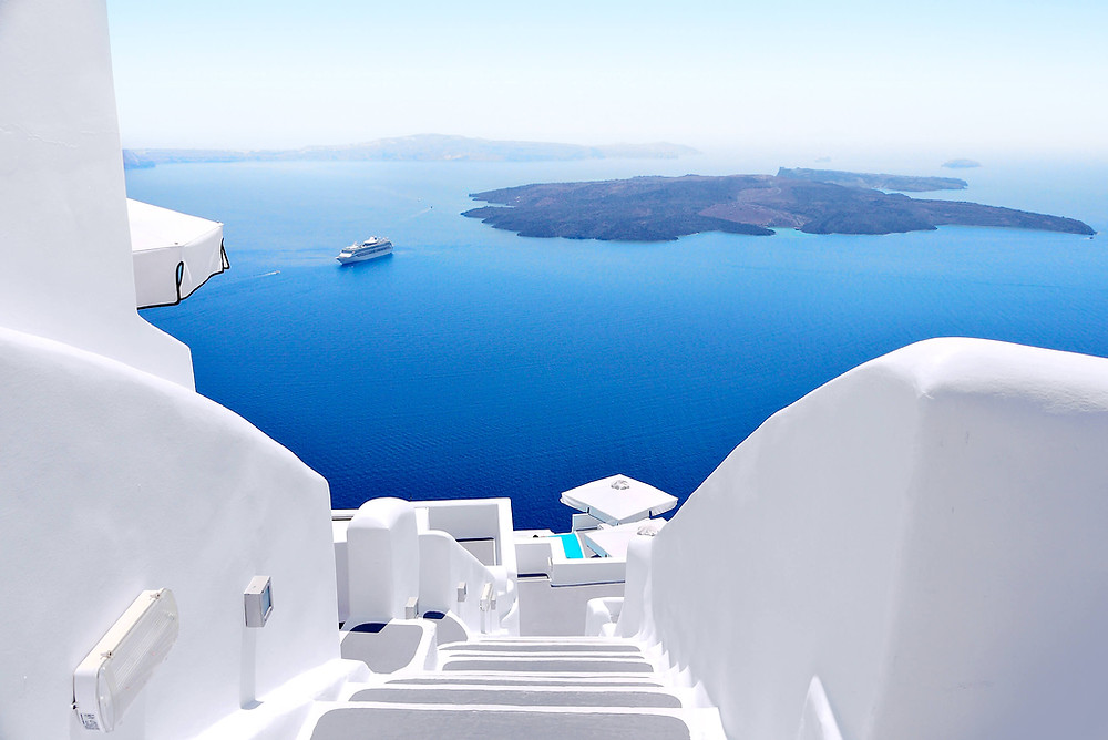 View down white staircases toward a deep blue sea with a distant island and a small cruise ship under clear sky