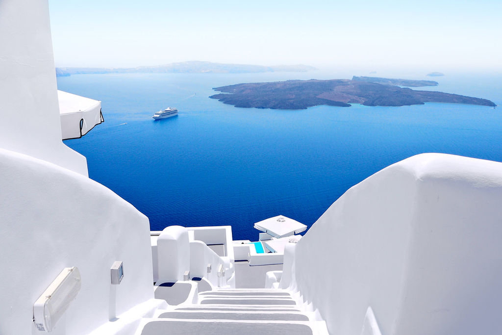 View down white staircases toward a deep blue sea with a distant island and a small cruise ship under clear sky